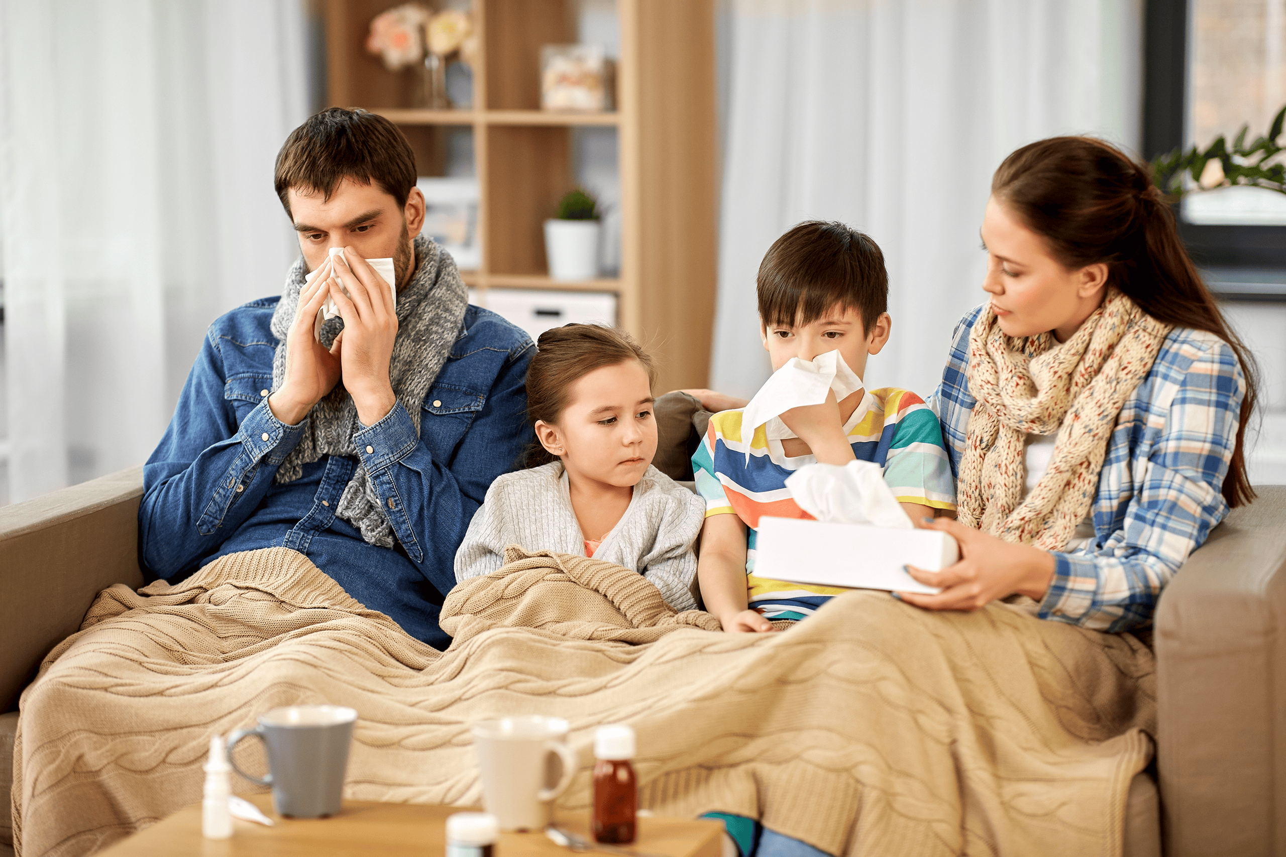 Familie mit Erkältungssymptomen auf dem Sofa, pharmazeutische Produkte im Vordergrund, pharmaselect.